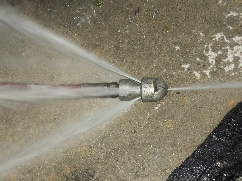 Close-up view inside a sewer pipe showing a high-pressure hydro jetting nozzle actively blasting multiple powerful streams of water forward and sideways to clear grease, debris, and buildup from the pipe walls.