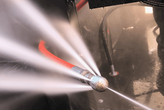 Inside a large, dark commercial sewer or grease interceptor: a red high-pressure hose with a spinning Warthog-style nozzle blasts intense white water jets in every direction, lighting up the wet interior as it scours away heavy grease and waste buildup.