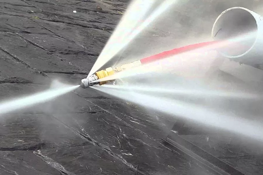 High-power hydro-jetting nozzle in full operation, blasting multiple intense white water jets in a wide fan pattern as it exits a large sewer pipe, demonstrating extreme cleaning force on a concrete surface.