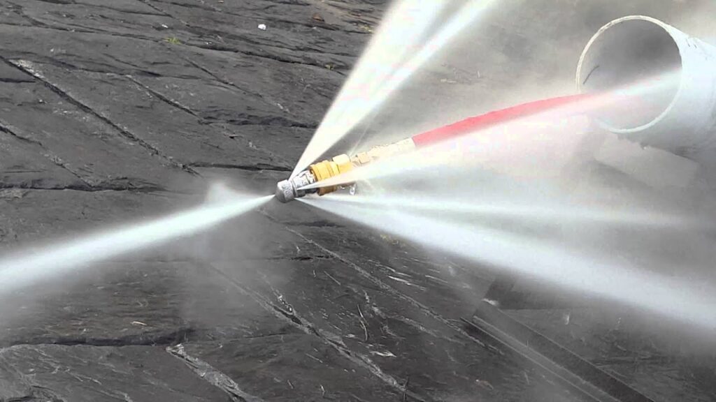 Close-up of a high-pressure sewer jetting nozzle blasting powerful rearward water jets while being pulled back through a large white PVC pipe, with a red jetter hose visible and water spraying forcefully onto surrounding dark soil.