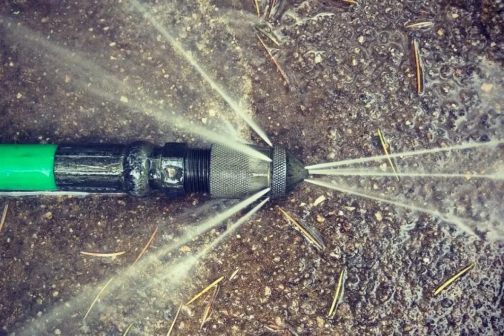 Close-up of a high-pressure sewer jetter nozzle actively blasting multiple powerful forward and backward water jets while lying on wet dirt, attached to a green-and-black jetter hose.