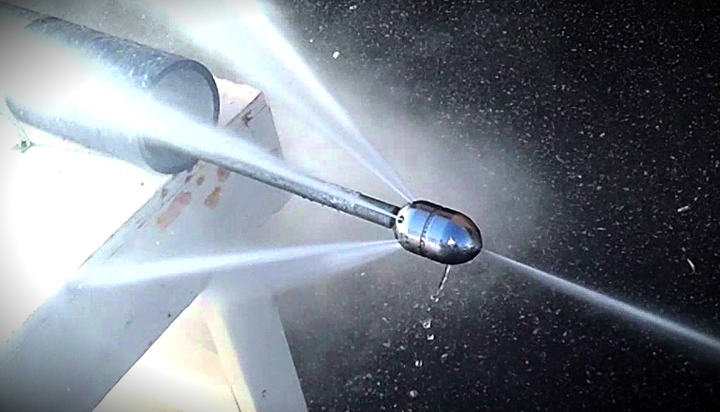 Close-up of a high-pressure hydro-jetting nozzle in action inside a dark sewer pipe, shooting intense forward and sideways water jets that light up the pipe with bright white streams as it powers through blockages.