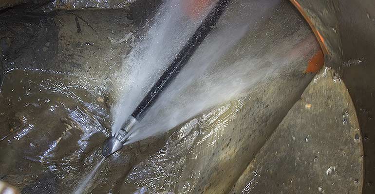 Inside a large sewer main: a high-pressure hydro-jetting nozzle blasts powerful streams of water against heavily encrusted grease and sludge buildup on the pipe walls, actively scouring and flushing debris downstream.