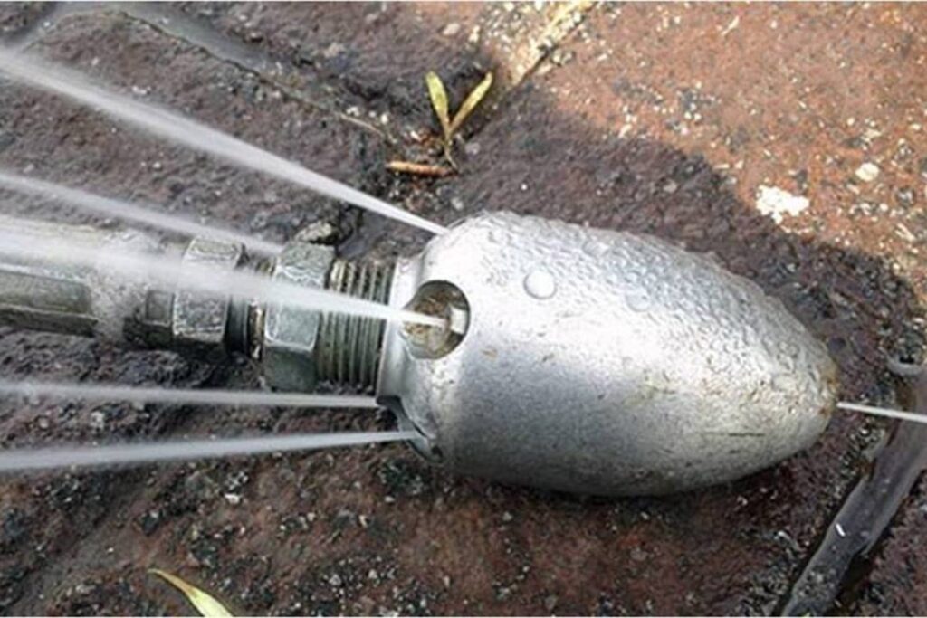 Close-up of a heavy-duty root-cutting hydro-jetting nozzle in action, with multiple high-pressure water jets shooting sideways and forward from a bullet-shaped metal head, designed specifically for shredding tree roots in sewer lines.