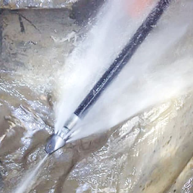 Close-up inside a sewer pipe of a high-pressure hydro-jetting nozzle blasting powerful forward and backward water jets, scouring heavy grease, sludge, and debris from the pipe walls during professional sewer cleaning.