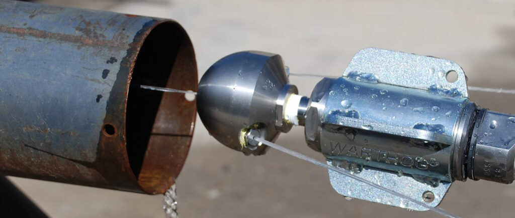 Close-up of a Warthog brand professional hydro-jetting nozzle attached to a high-pressure hose, positioned at the opening of a rusted metal pipe, with water droplets on the polished stainless-steel head ready for sewer cleaning.