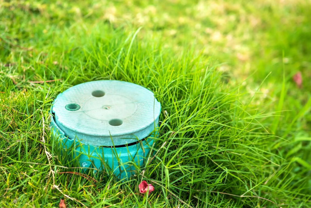 A round, turquoise-green plastic irrigation valve box with a matching lid set flush into a lush green lawn, providing access to underground sprinkler control valves.