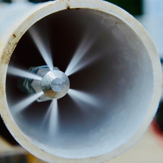Inside a clean white PVC drain pipe: a spinning hydro-jetting nozzle shoots bright white high-pressure water jets in a perfect 360-degree starburst pattern as it travels forward, scouring the pipe walls spotless.