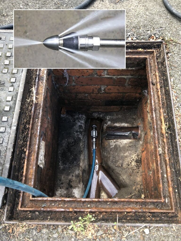 Open brick sewer cleanout/manhole with a blue high-pressure hydro-jetting hose inserted into the main line; inset photo shows the spinning nozzle blasting powerful water jets in all directions.