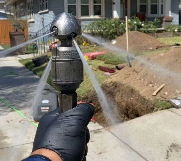 Gloved hand holding a Warthog hydro-jetting nozzle blasting powerful water jets in multiple directions during a residential sewer cleaning job, with a suburban house, front yard, and open sewer cleanout visible in the background.