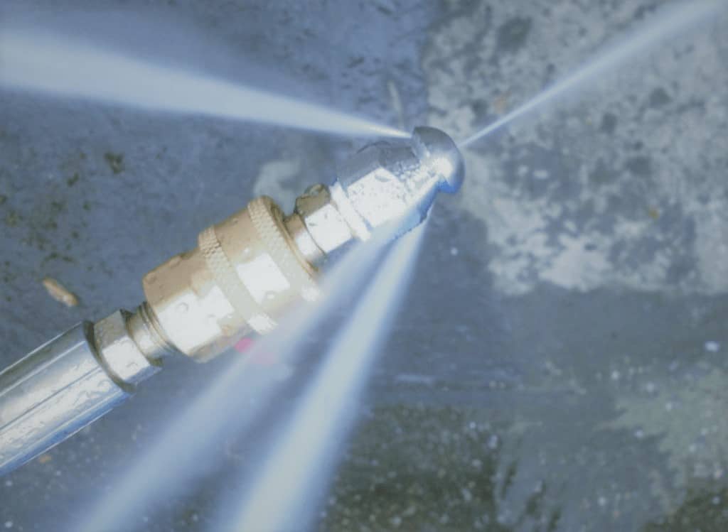 Dramatic close-up of a professional hydro-jetting nozzle in full blast, shooting four razor-sharp high-pressure water jets in a symmetrical pattern against a dark sewer floor, showcasing raw 4,000 PSI cleaning power.