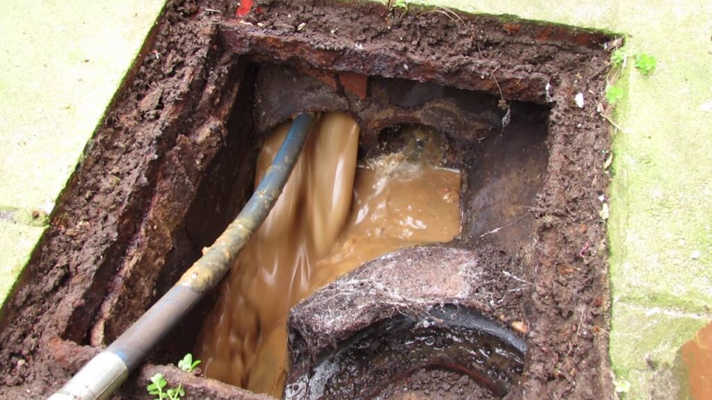 Close-up of a severely clogged sewer cleanout overflowing with thick brown wastewater and sewage as a drain snake cable is pushed into the flooded access point, showing a major main line blockage.