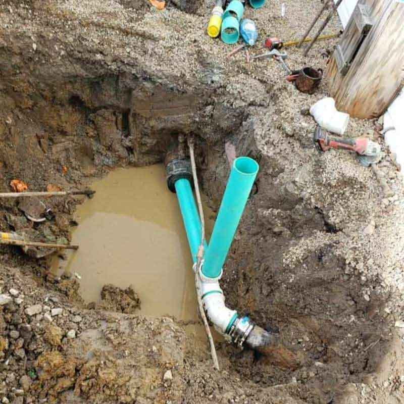 Open excavation showing a broken and misaligned underground sewer line with standing wastewater; new turquoise PVC pipes and fittings are partially installed as part of the repair, surrounded by muddy soil and scattered tools.