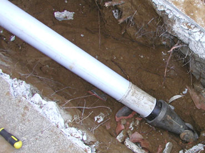 White PVC sewer pipe emerging from under a cracked concrete slab, connected to an old cast-iron or clay pipe stub with a black rubber Fernco shielded coupling in a muddy excavation next to a building foundation.