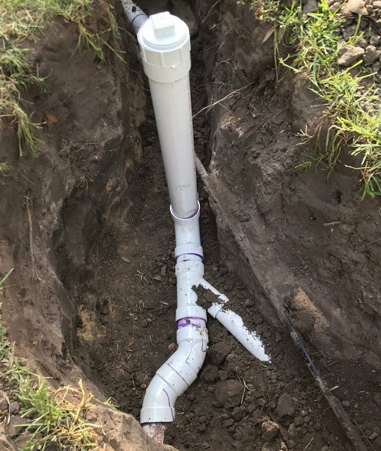 White PVC two-way sewer cleanout riser with threaded cap exposed in a shallow hand-dug hole, showing a long-sweep 90-degree elbow and horizontal run freshly glued with purple primer visible on the joints.