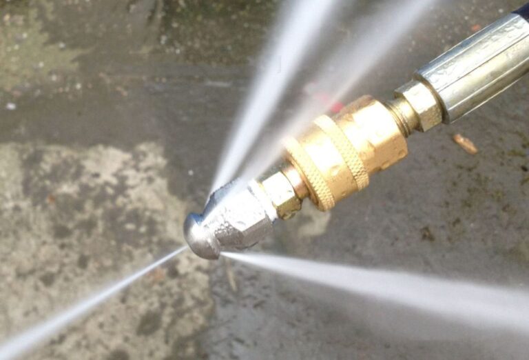 Close-up of a high-pressure sewer jetting nozzle in full operation, spraying multiple intense white water jets in a wide fan pattern from a polished stainless-steel head attached to a brass quick-connect fitting.